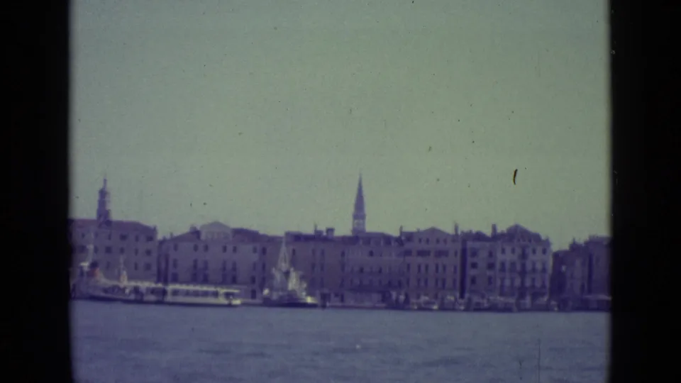 1962 Venice canal scene with low boats, waterfront buildings, and a tall spire visible beyond the roofline.