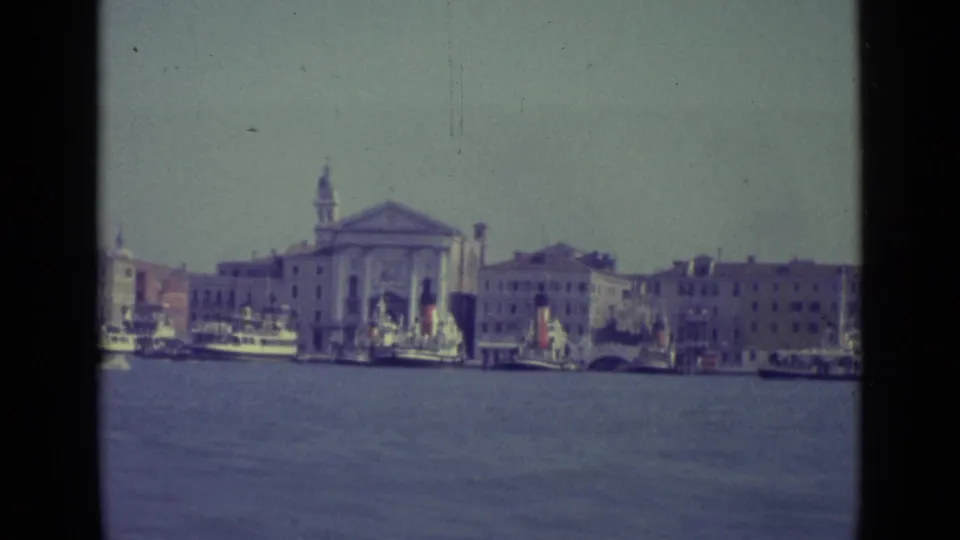 1962 8mm Venice view from the water showing a church facade, bell tower, moored boats, and waterfront buildings.
