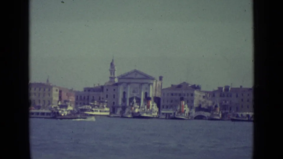 1962 Venice waterfront seen from a boat, with lagoon water, docked vessels, pale buildings, and a church bell tower.