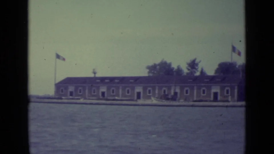 1962 8mm view of a low island building in Venice, with flags, shoreline, trees, and open lagoon water in front.