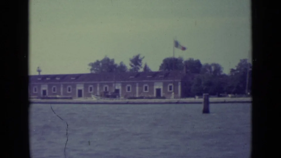 1962 Venice lagoon view of a low island structure with flags flying, trees behind it, and a channel marker in the water.