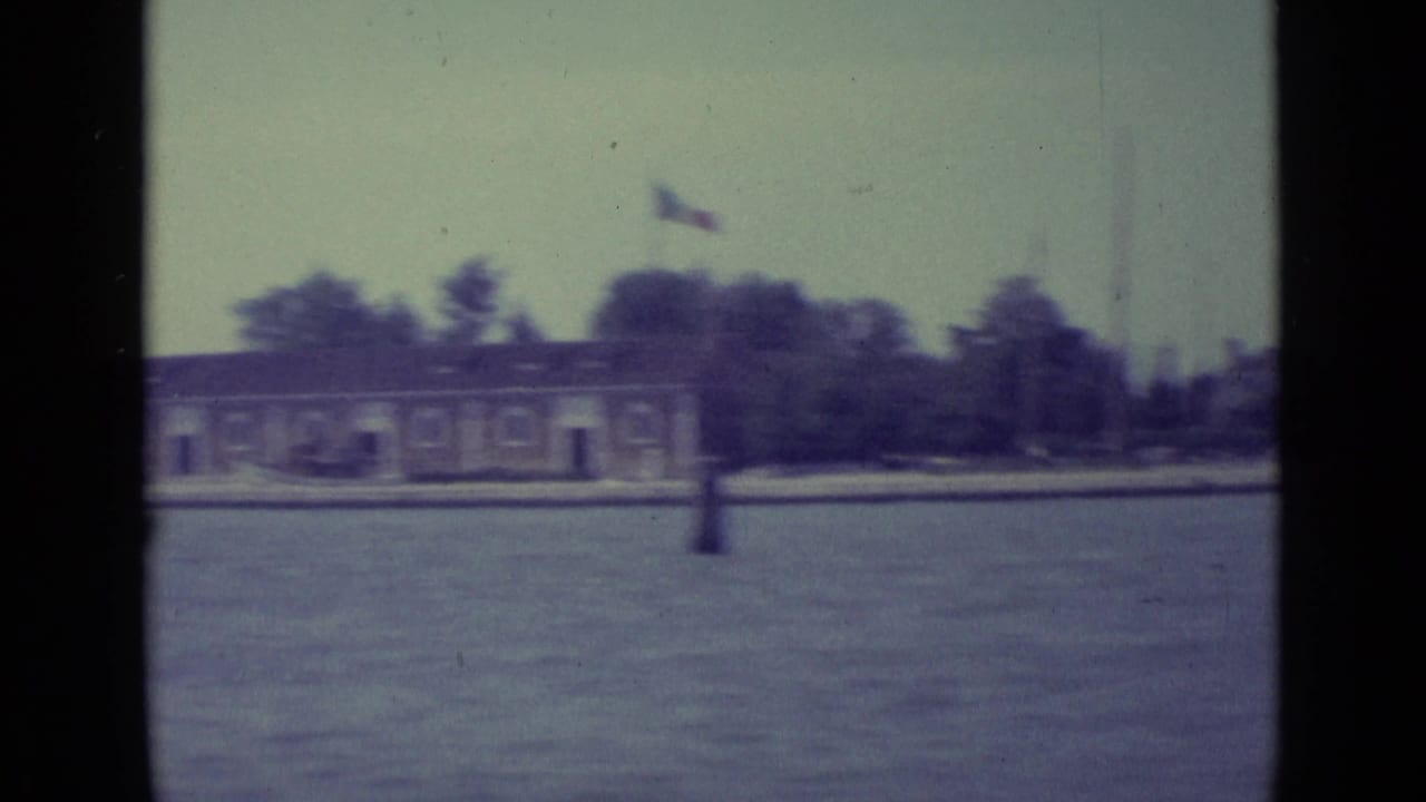 Distant shoreline and low building seen across Venetian water through dark film-frame borders.