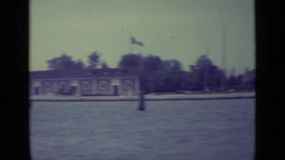 1962 Venice home-movie frame showing a distant shoreline, low building, trees, flag, water, and heavy 8mm frame borders.