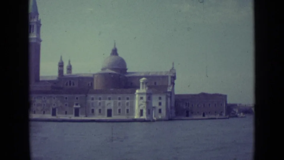 The pan crops the tall tower at the edge while showing the domed Venice waterfront building and adjacent low structure across the lagoon.