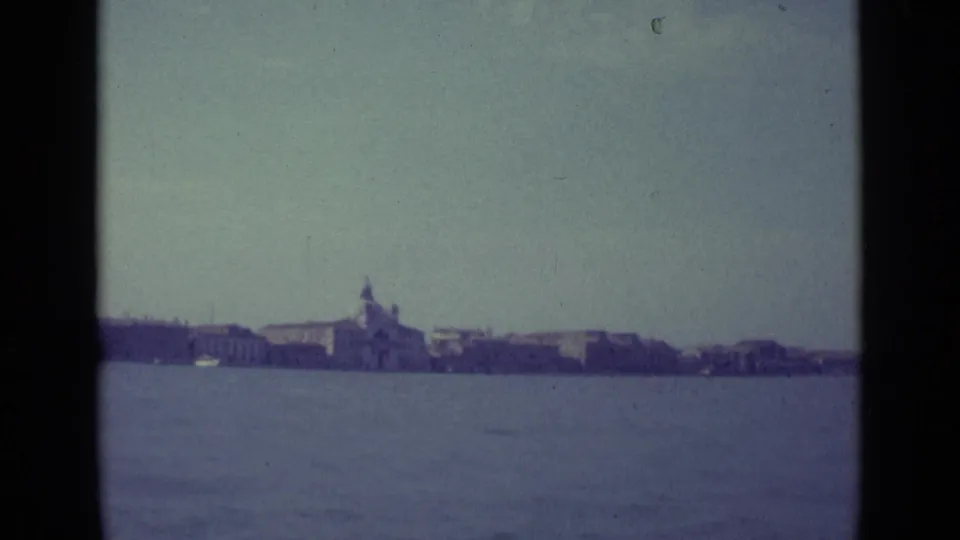 Wide 1962 home-movie view across the Venice lagoon toward a distant shoreline with low buildings and a small central tower form.