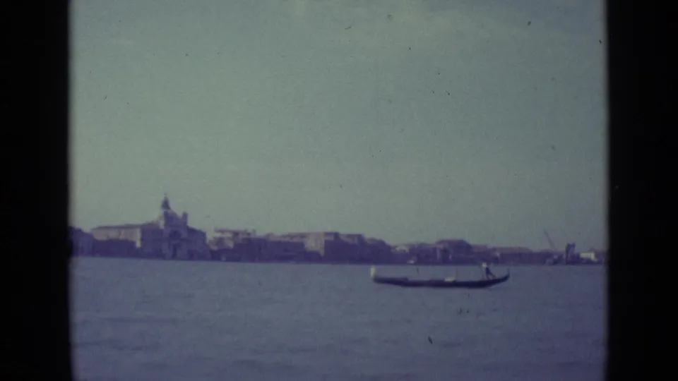 1962 home-movie frame of a gondola crossing the Venice lagoon, with the city shoreline and domed church in the background.