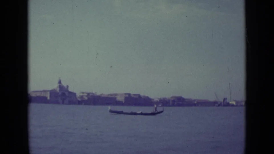 1962 8mm Venice lagoon view with a gondola in mid-frame and a distant shoreline of buildings and a domed church.