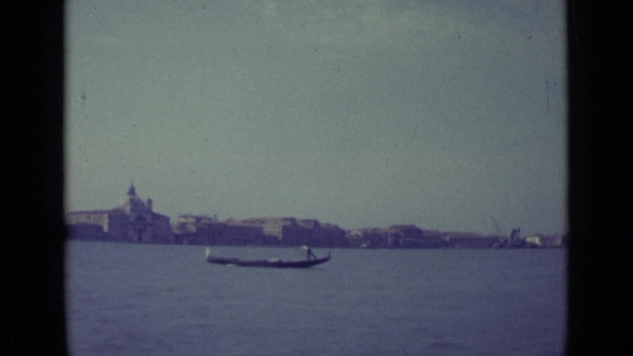 1962 8mm view of Venice, Italy, with a gondola moving across lagoon water before a hazy waterfront skyline.