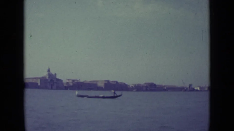 1962 8mm view of Venice, Italy, with a gondola moving across lagoon water before a hazy waterfront skyline.