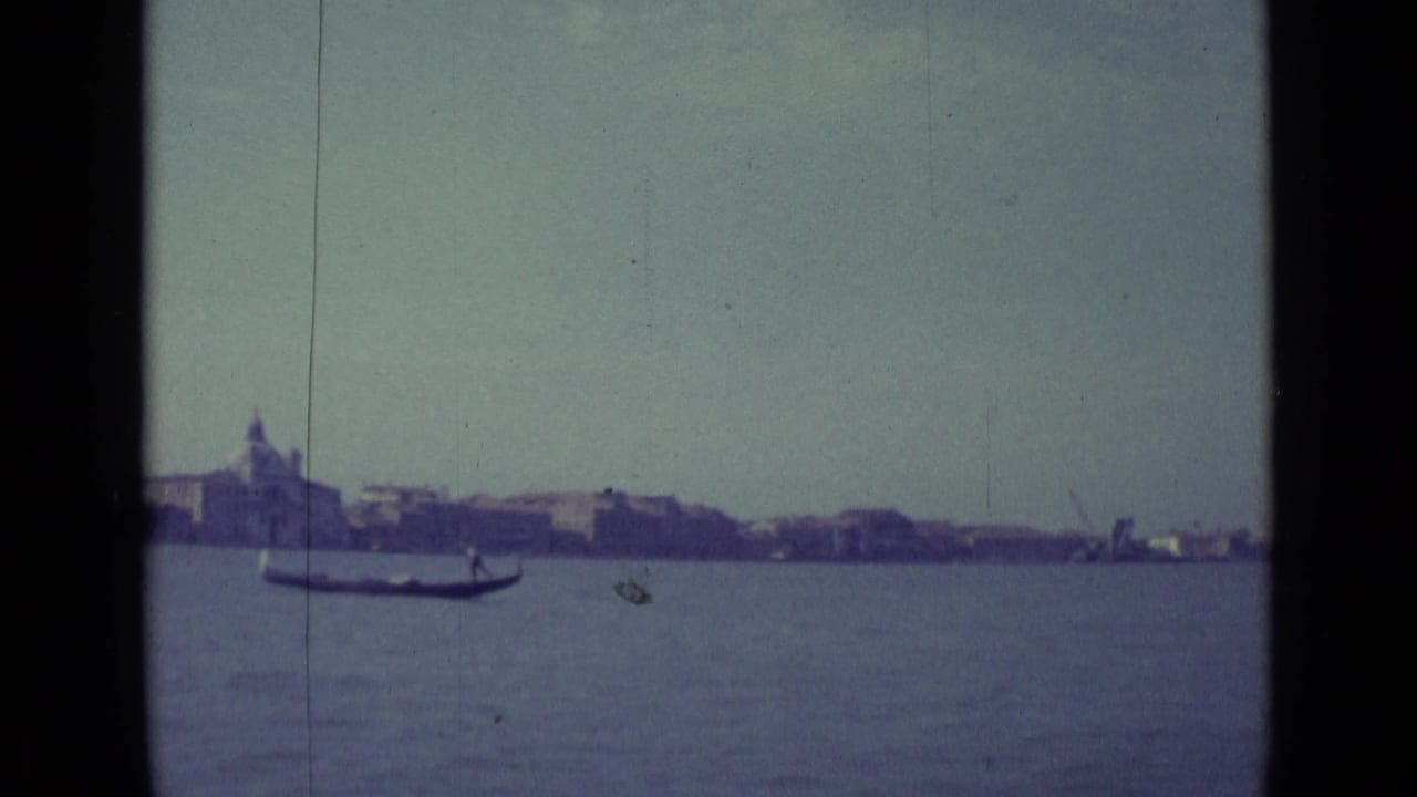 Gondola on calm water before a distant waterfront skyline under hazy sky.