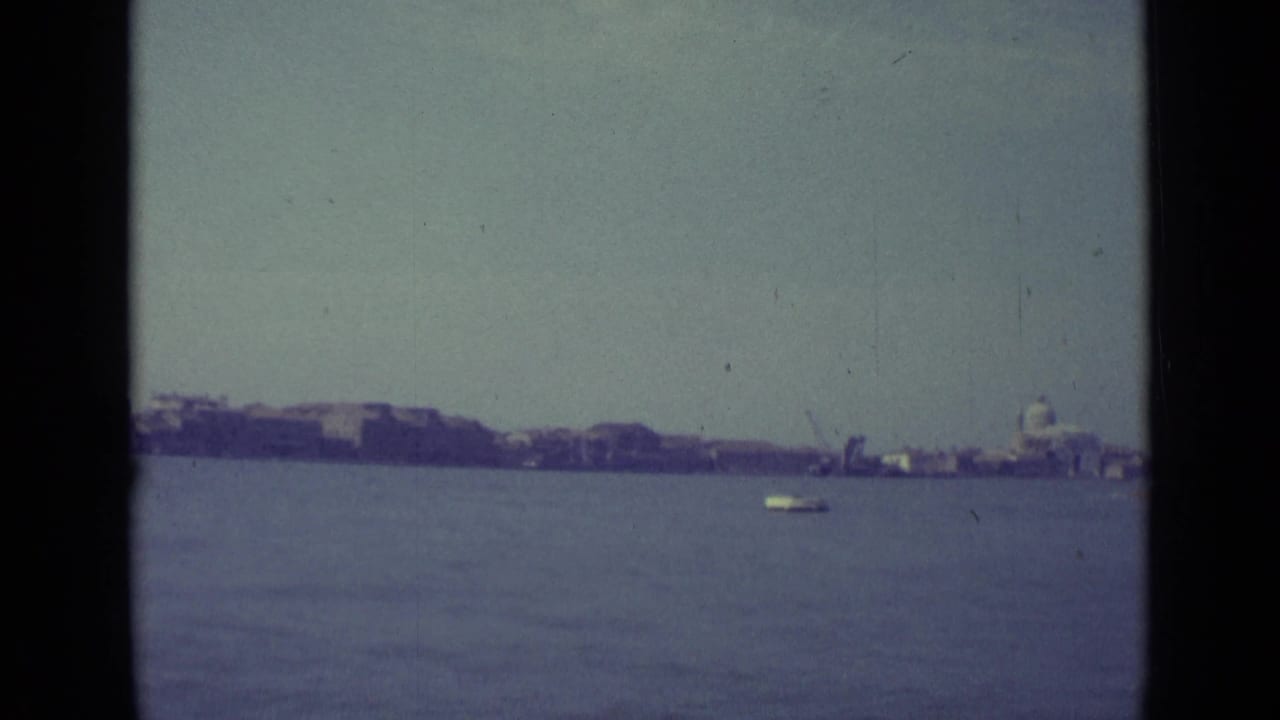 1962 Venice home-movie frame showing calm lagoon water, a small boat, and a distant domed building along the shoreline.