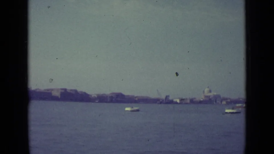 1962 Venice waterfront view from a boat, with open lagoon water, a small craft, and a distant dome on the skyline.