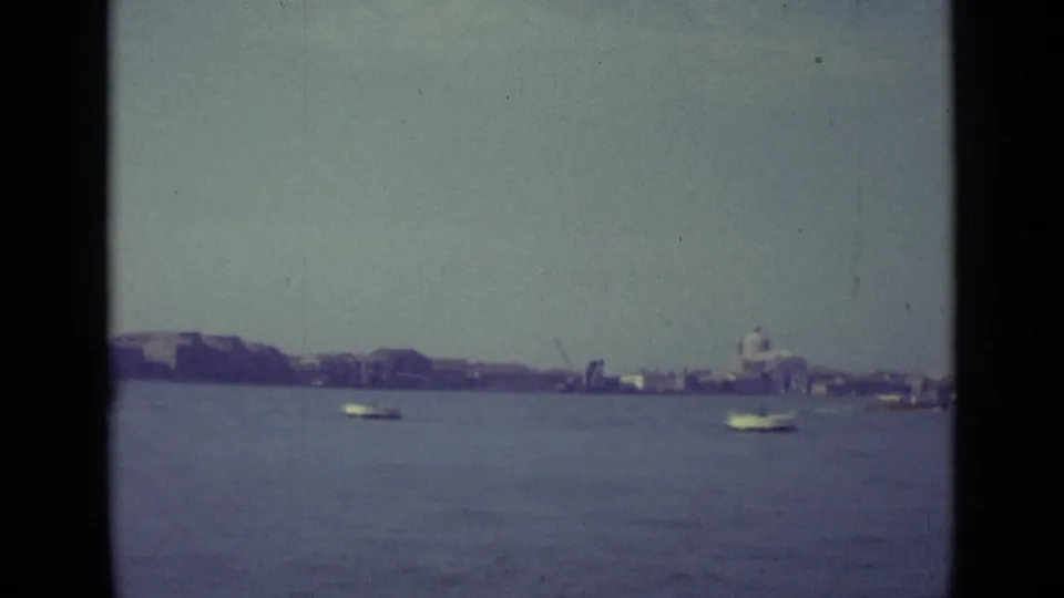 1962 Venice lagoon frame with multiple small boats on calm water and a distant domed building near the waterfront.