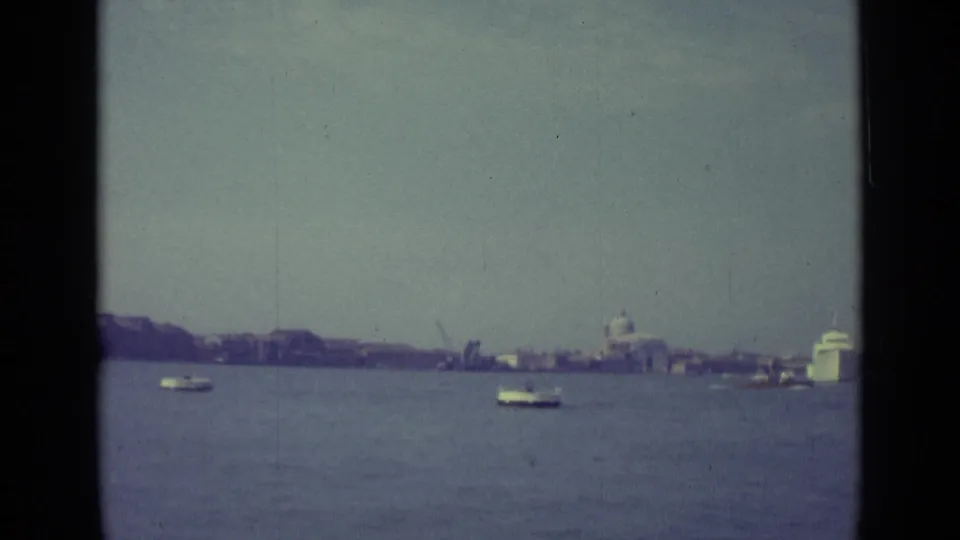 1962 Venice home-movie view across the lagoon, with small boats, a distant skyline, and a bright waterfront structure at right.