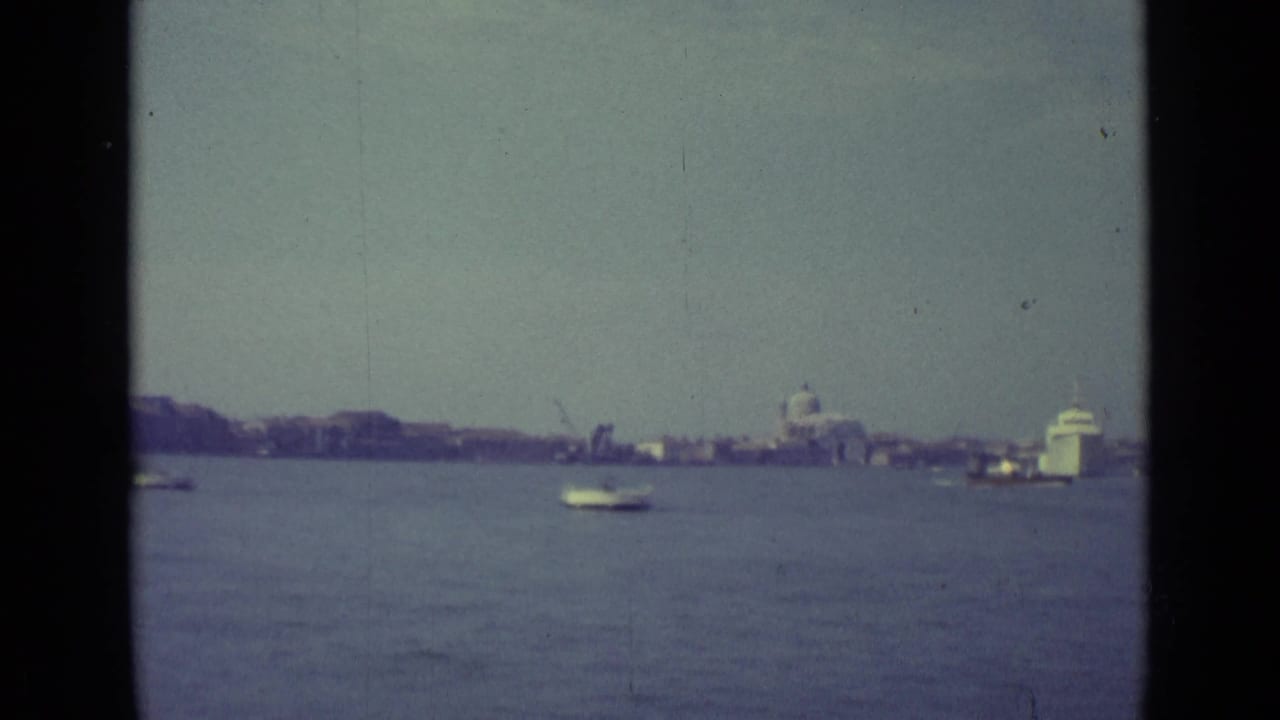 1962 8mm Venice view from the water, with small boats, a distant domed building, waterfront structures, and visible film-gate edges.