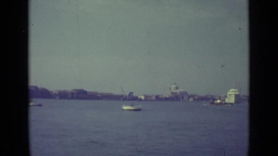 1962 8mm Venice view from the water, with small boats, a distant domed building, waterfront structures, and visible film-gate edges.