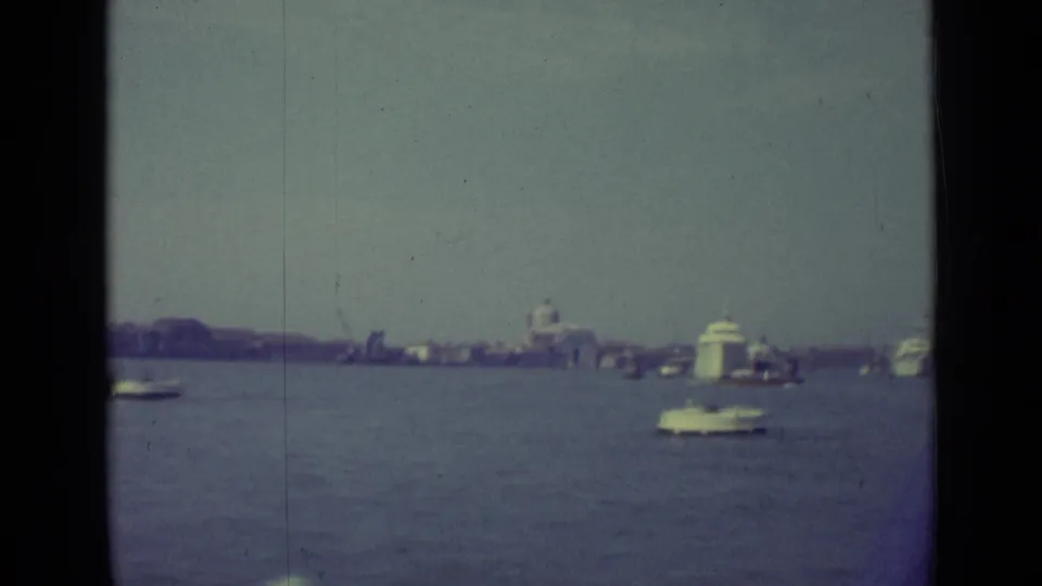 The 1962 home-movie frame shows a white boat in the foreground water with Venice waterfront buildings and a dome in the distance.