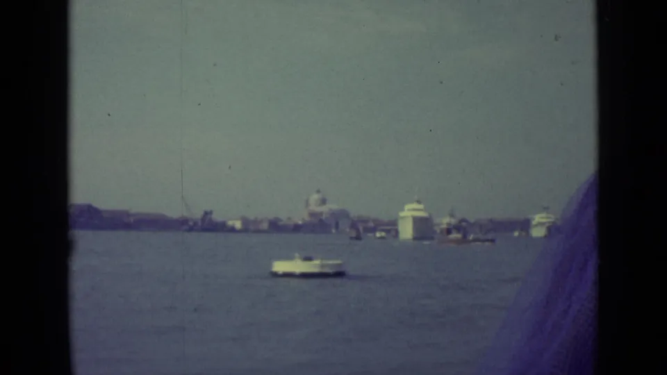 A handheld 8mm frame from Venice in 1962 shows open water, several boats, distant buildings, and a dark film or obstruction edge at right.