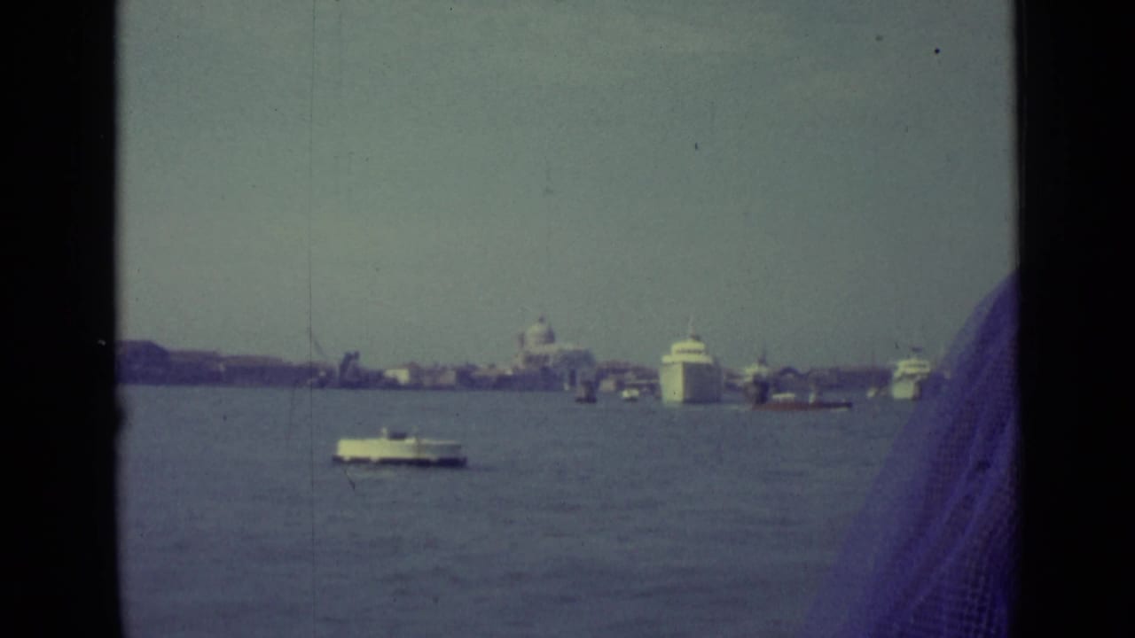 1962 Venice canal view from a moving boat, with water traffic, distant domes, dark window framing, and a red MONACO building at right.