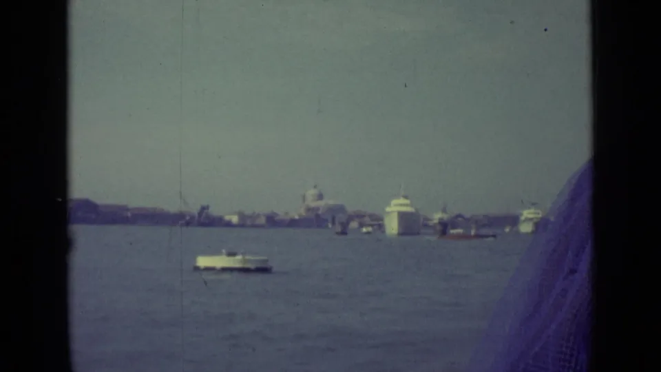1962 Venice canal view from a moving boat, with water traffic, distant domes, dark window framing, and a red MONACO building at right.