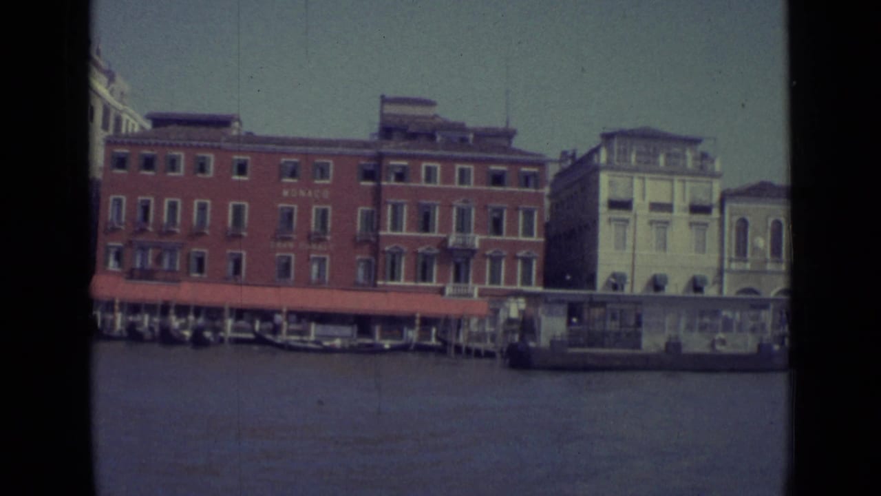 Red waterfront building with MONACO signage and red awning seen across the canal beside pale buildings.