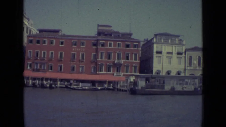 Venice waterfront in 1962 showing a red building marked MONACO, red awning, moored gondolas, and neighboring canal-side architecture.