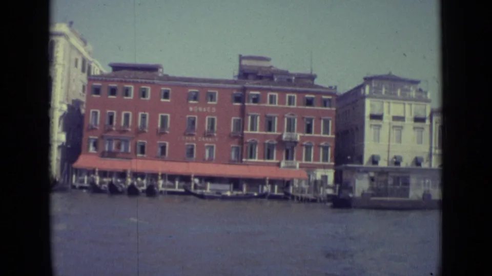 1962 Venice canal frontage with a red MONACO building, red awning, gondolas at the landing, and pale historic buildings alongside.