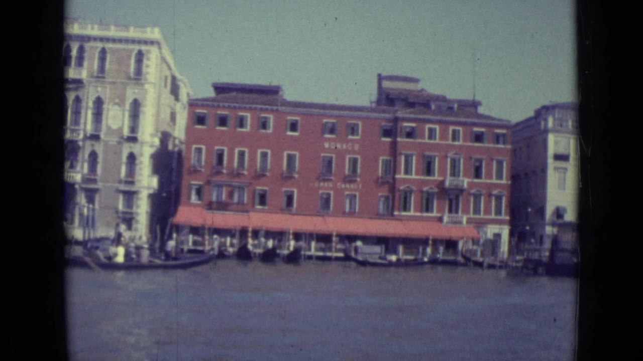 Grand Canal view with a red brick waterfront building, pale palazzo, awnings, boats, and gondolas.