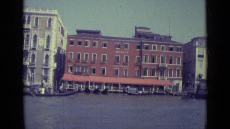 1962 Venice Grand Canal view showing a red brick building marked MONACO beside a pale palazzo, with gondolas and small boats on the water.