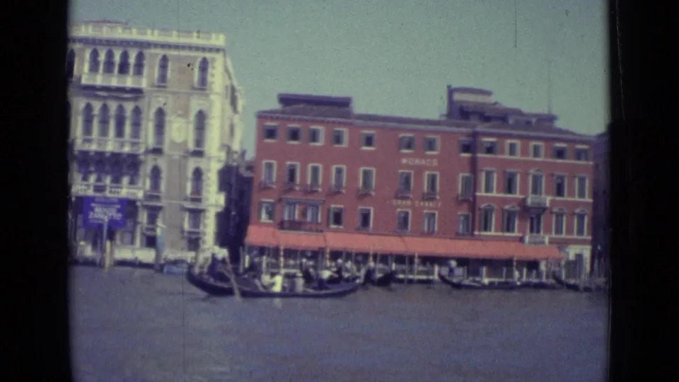 1962 Venice canal scene with gondolas crossing before a pale arched palazzo and the red MONACO waterfront building with awnings.