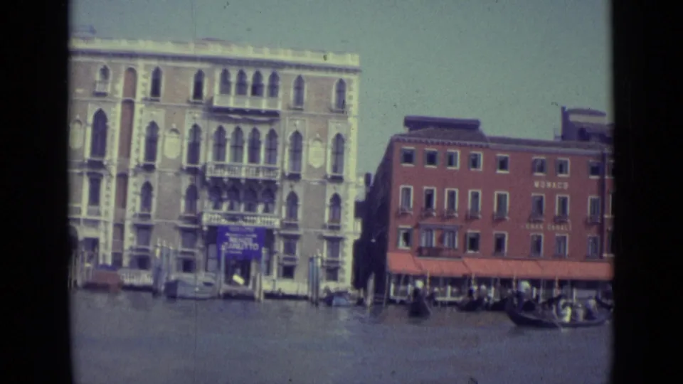 1962 Venice Grand Canal shot showing a pale arched palazzo, the red MONACO building, waterfront awnings, and gondolas near frame edge.