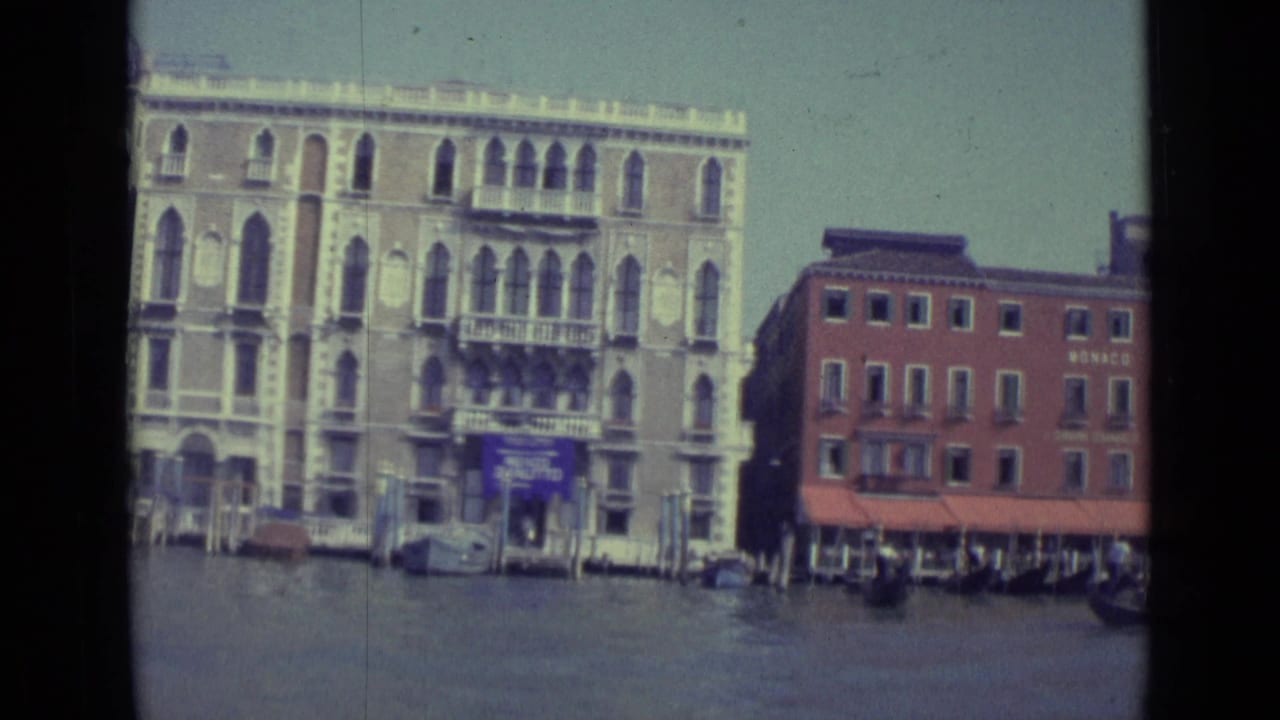 1962 Venice canal view with arched historic facades, balconies, mooring poles, water, and a red MONACO-marked building.