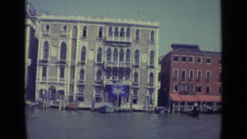 1962 Venice canal frontage with pale arched architecture, balcony railings, mooring poles, and the red MONACO-marked building.