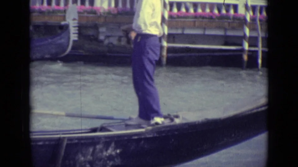1962 Venice detail of a gondolier in a light shirt and dark trousers standing on a black gondola near a canal-side facade.