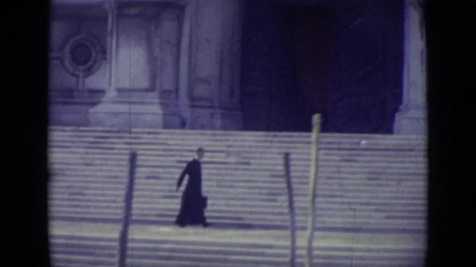 1962 Venice 8mm frame showing a priest in a dark cassock crossing broad stone steps near a large cathedral-like facade.