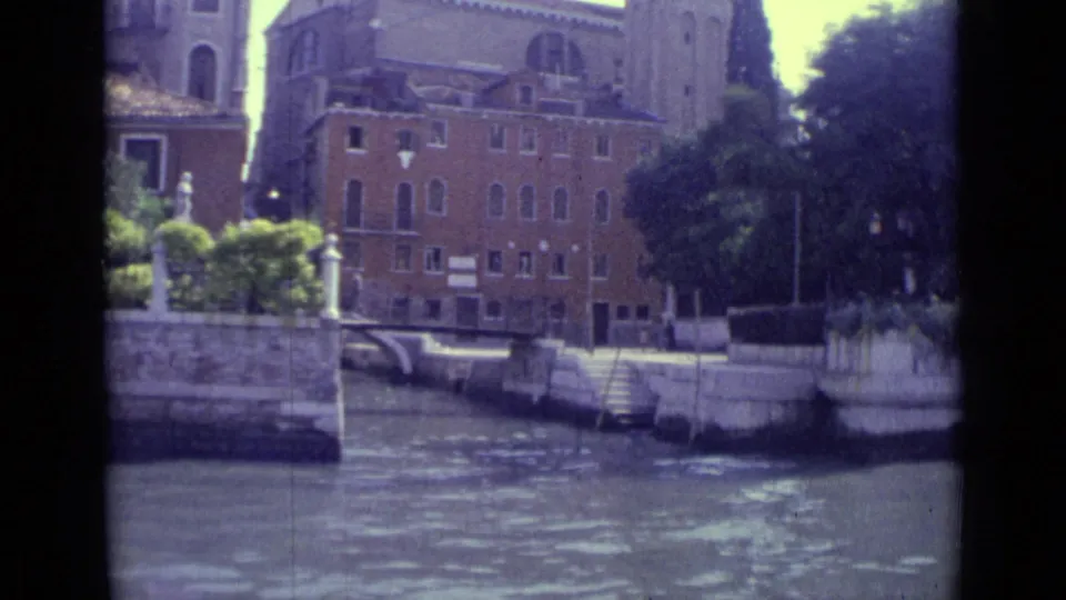 1962 Venice canal-side view with brick buildings, moored boats, stone steps, and trees along the water.