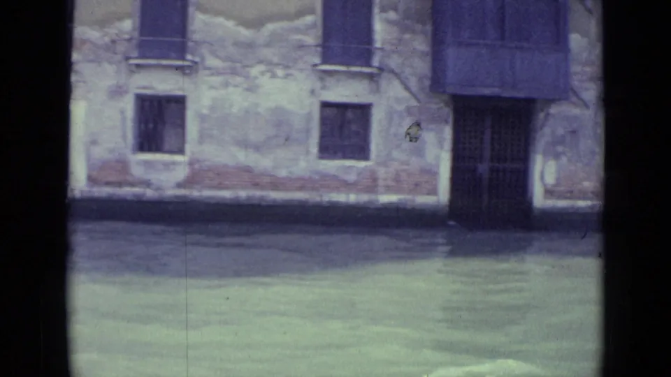 1962 Venice canal footage showing water, worn plaster, shuttered windows, and a dark recessed doorway at the building edge.