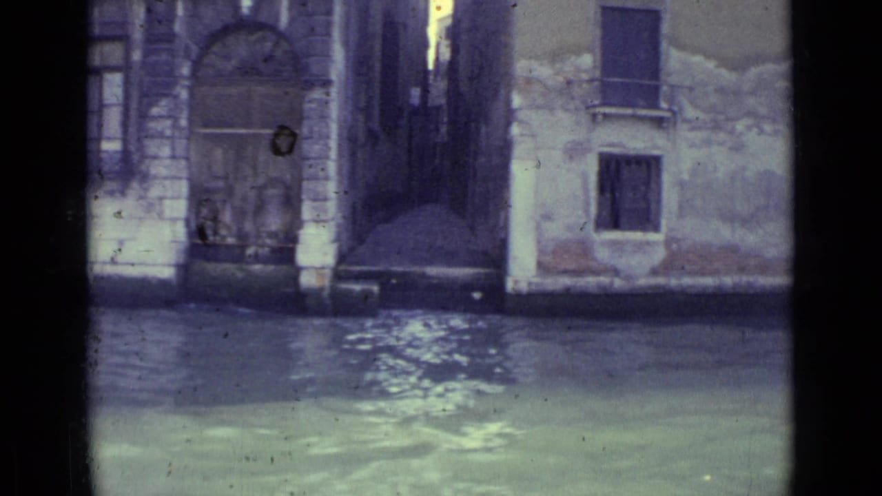 1962 Venice 8mm frame showing canal water below weathered masonry facades, arched doors and small shuttered windows.