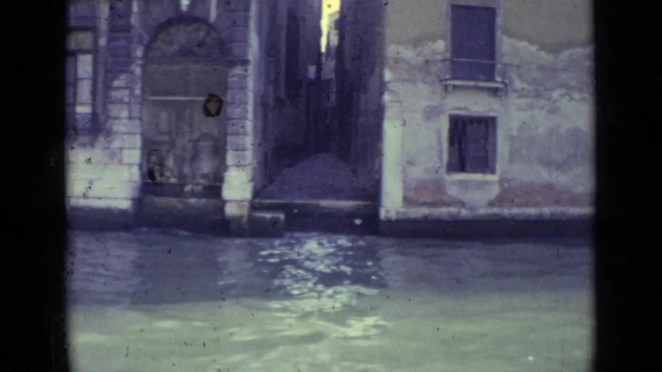 1962 Venice 8mm frame showing canal water below weathered masonry facades, arched doors and small shuttered windows.
