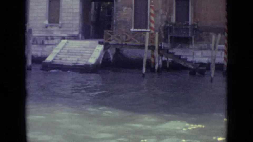 Venice 8mm frame showing steps down to the canal, a small dock structure, striped mooring poles and rippling water.