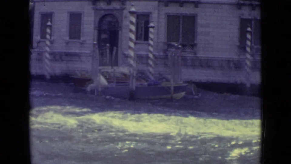 1962 Venice canal view with rippling water, a stone facade, shuttered windows, and striped mooring poles.