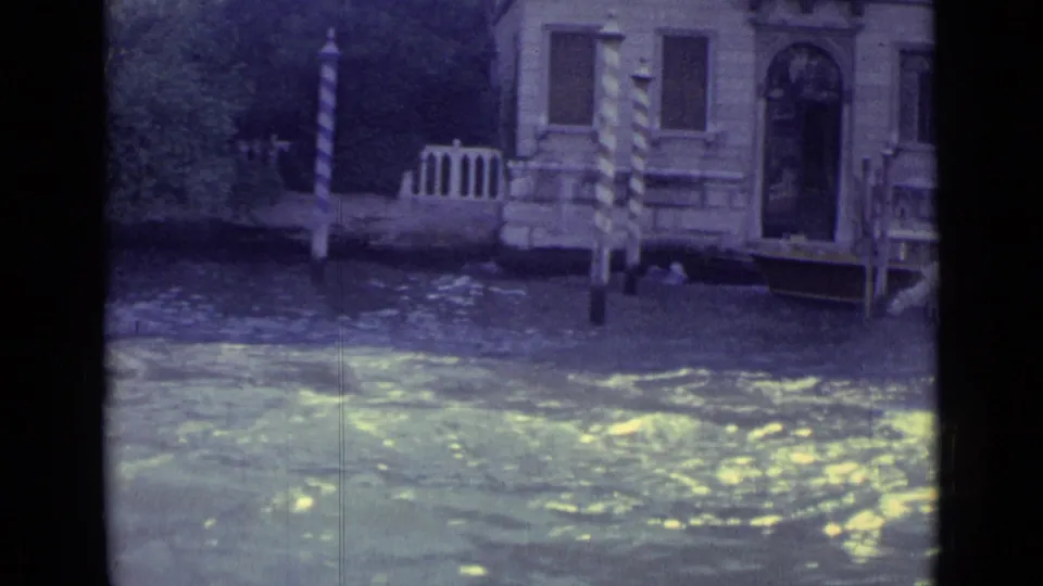 Visitor-shot 1962 Venice canal view with water, a building entrance, and striped mooring poles near a small landing.