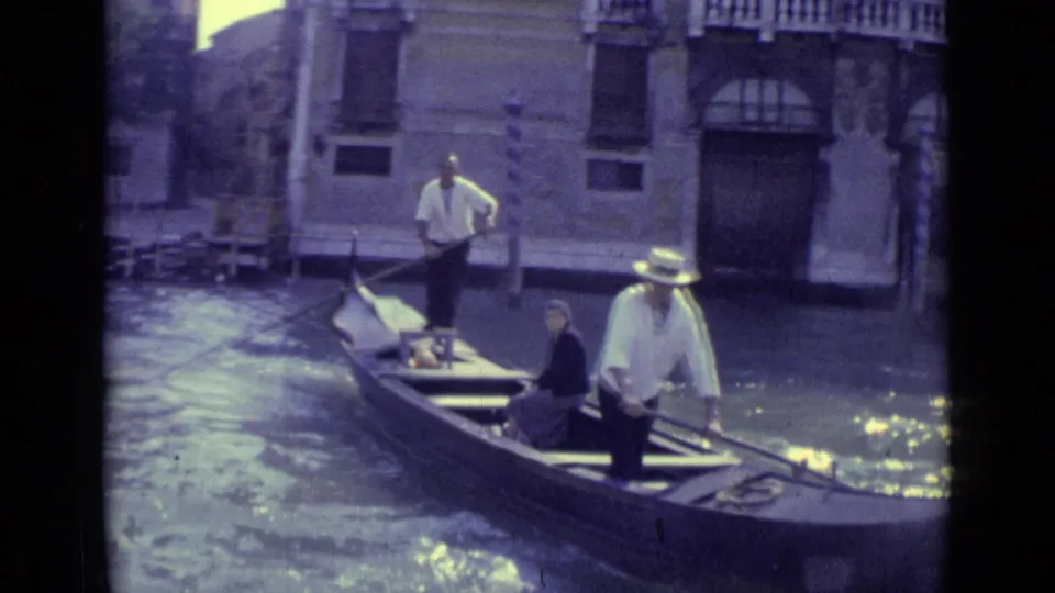 A 1962 Venice gondola passes on the canal, with a gondolier standing and a passenger seated near stone buildings.