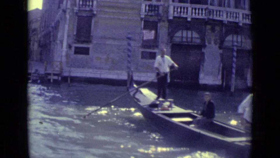 1962 Venice 8mm frame showing a gondola passing a stone canal building with a gondolier standing at the stern and a passenger seated aboard.