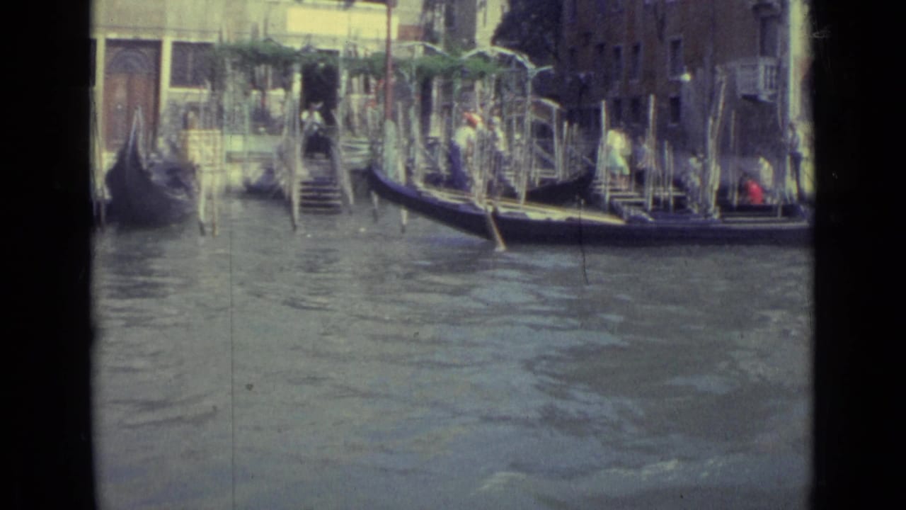 Gondola near canal steps with mooring poles, dockside figures, and waterfront buildings.