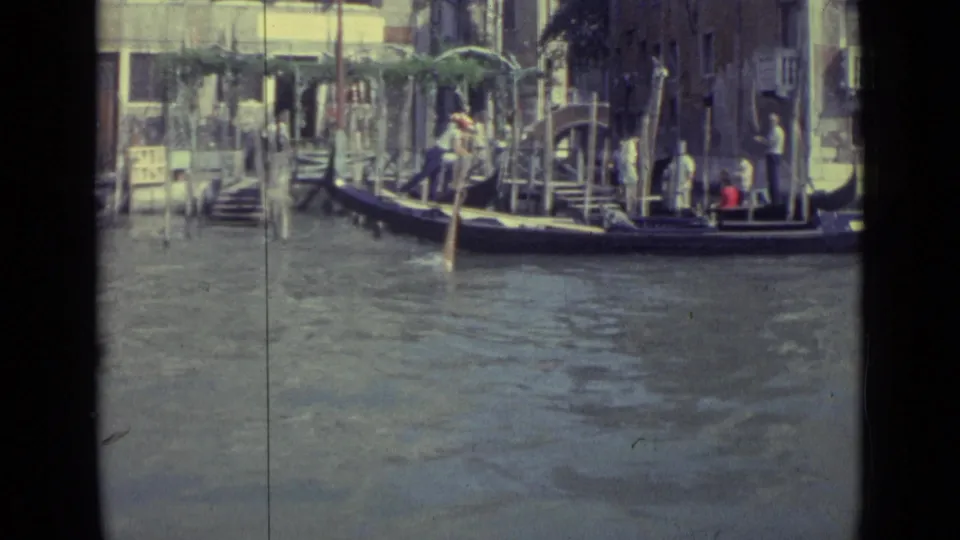 1962 Venice 8mm frame showing a gondola gliding past dock steps and clustered mooring poles, with people visible along the canal edge.