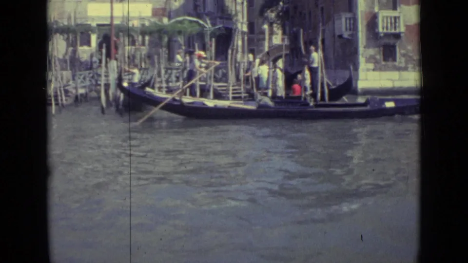1962 Venice frame showing a long gondola with a standing gondolier and seated passengers beside dock steps, mooring poles, and a canal facade.