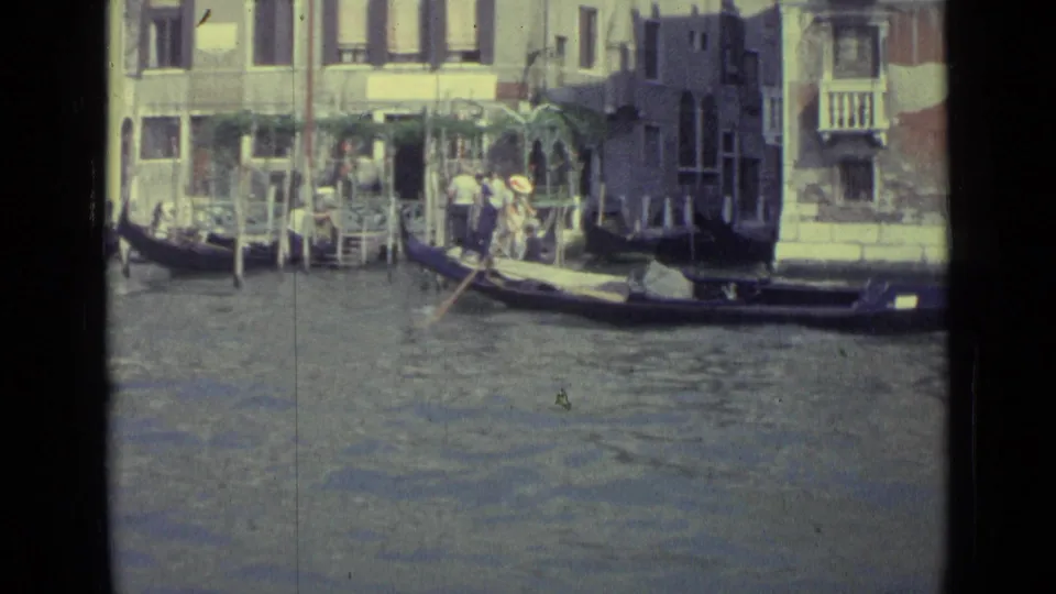 1962 Venice canal view with a gondola, gondolier, passengers, waterfront facades, balconies, and mooring poles.