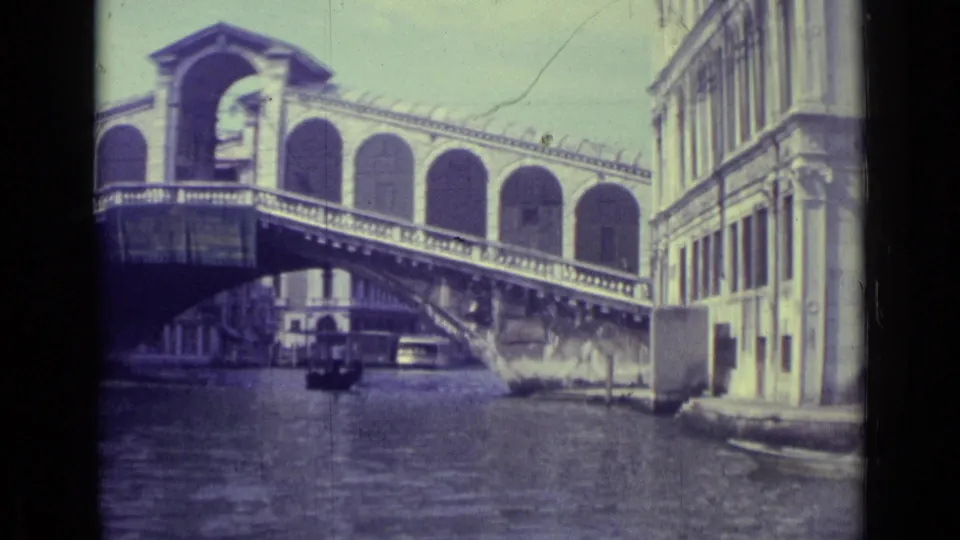 1962 8mm Venice footage frames the Rialto Bridge from the canal, with a boat below and tall waterfront buildings to the right.