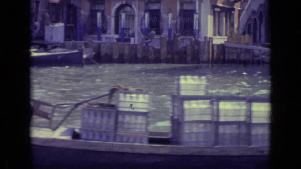 1962 Venice canal footage shows a boat loaded with crates passing in front of weathered facades and wooden mooring posts.